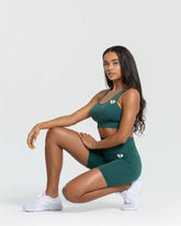 امرأة ترتدي حمالة صدر رياضية خضراء، شورت Power Seamless لركوب الدراجات | لون الغابة الأخضر، وحذاء رياضي أبيض يجلس على الأرض. اسم العلامة التجارية: أفضل النساء